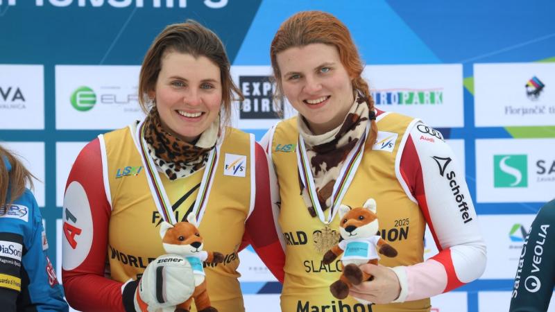 Two female athletes wearing a gold bib smile on the podium